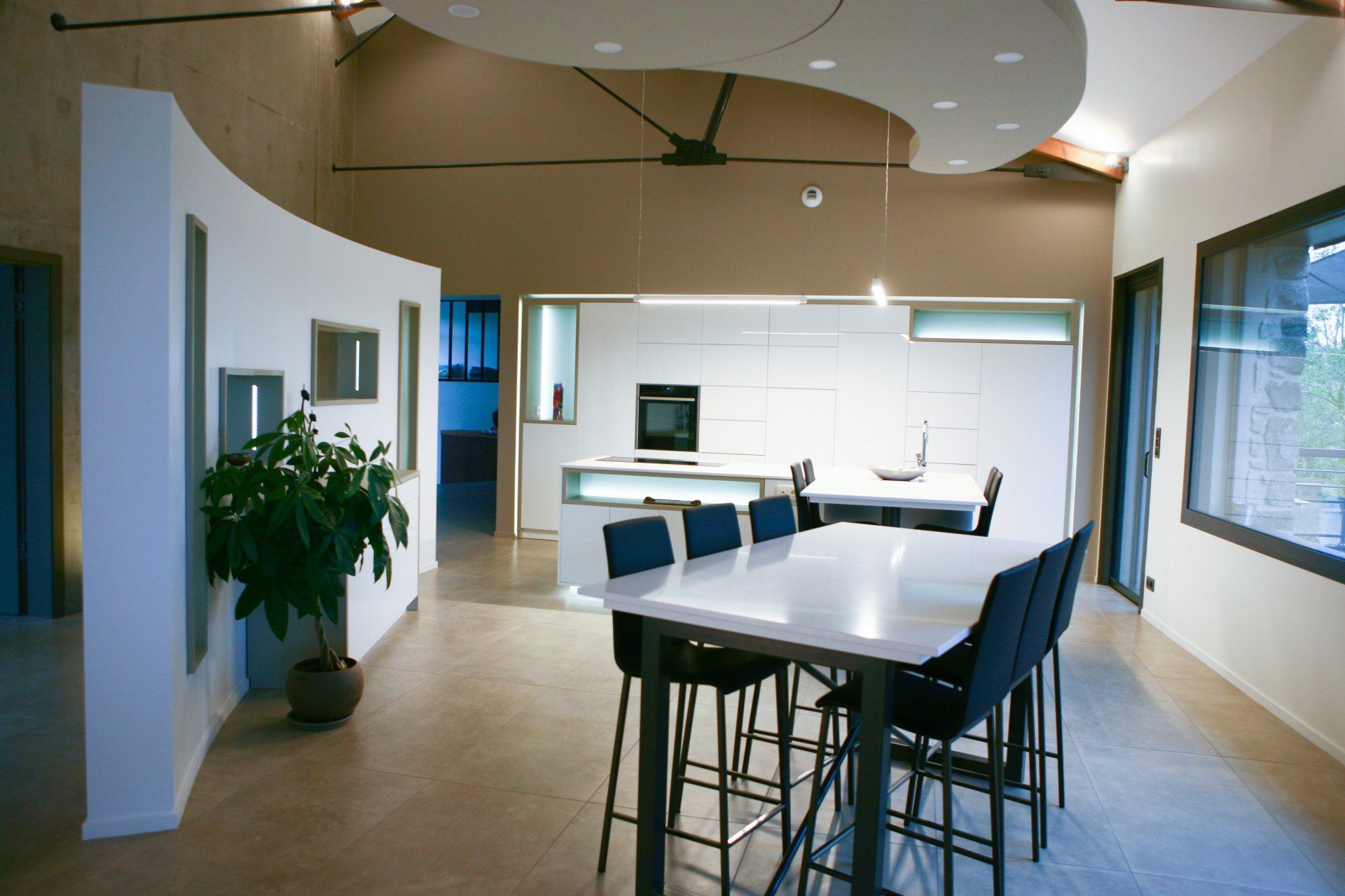 Une salle à manger moderne avec une table blanche, des chaises bleu foncé et un mur incurvé sur la gauche, menant à une cuisine ouverte.