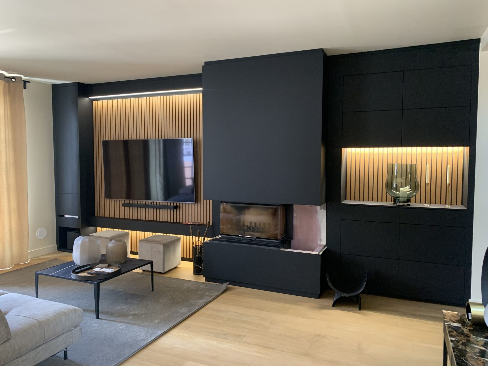 Un salon avec un meuble mural noir intégrant une cheminée, une télévision, des accents de lattes de bois et un éclairage chaleureux.