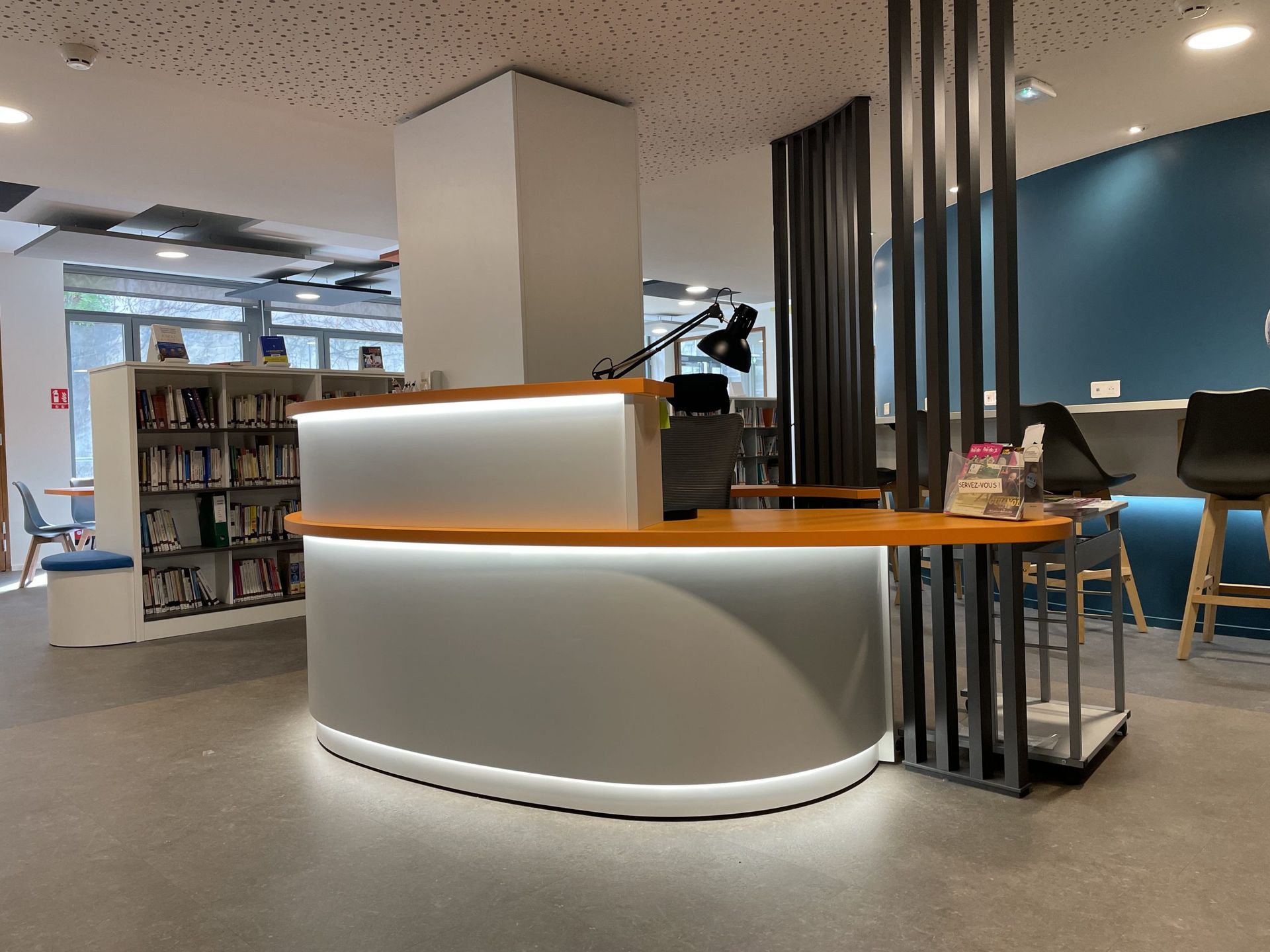 Un comptoir d'accueil moderne et incurvé avec éclairage sous le comptoir, dans une bibliothèque contemporaine, avec des étagères et des tabourets de bar.