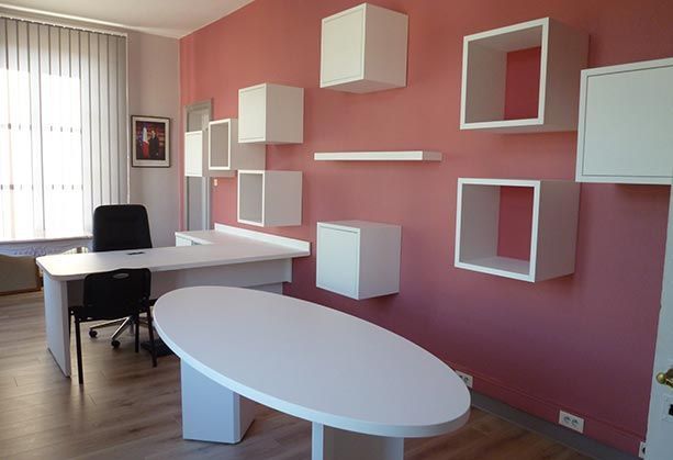 Un bureau avec un bureau ovale blanc, un bureau d'angle, deux chaises et des étagères cubiques blanches sur un mur rose.