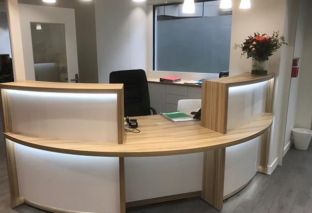 Un comptoir d'accueil moderne en bois courbé, avec des panneaux lumineux et un bouquet de fleurs, dans un hall d'immeuble de bureaux baigné de lumière.