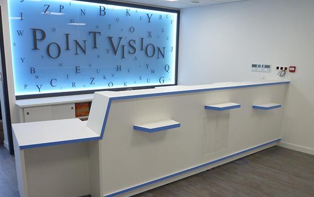 Un comptoir d'accueil blanc moderne avec des touches de bleu se trouve devant une enseigne murale rétroéclairée sur laquelle on peut lire « POINT VISION ».