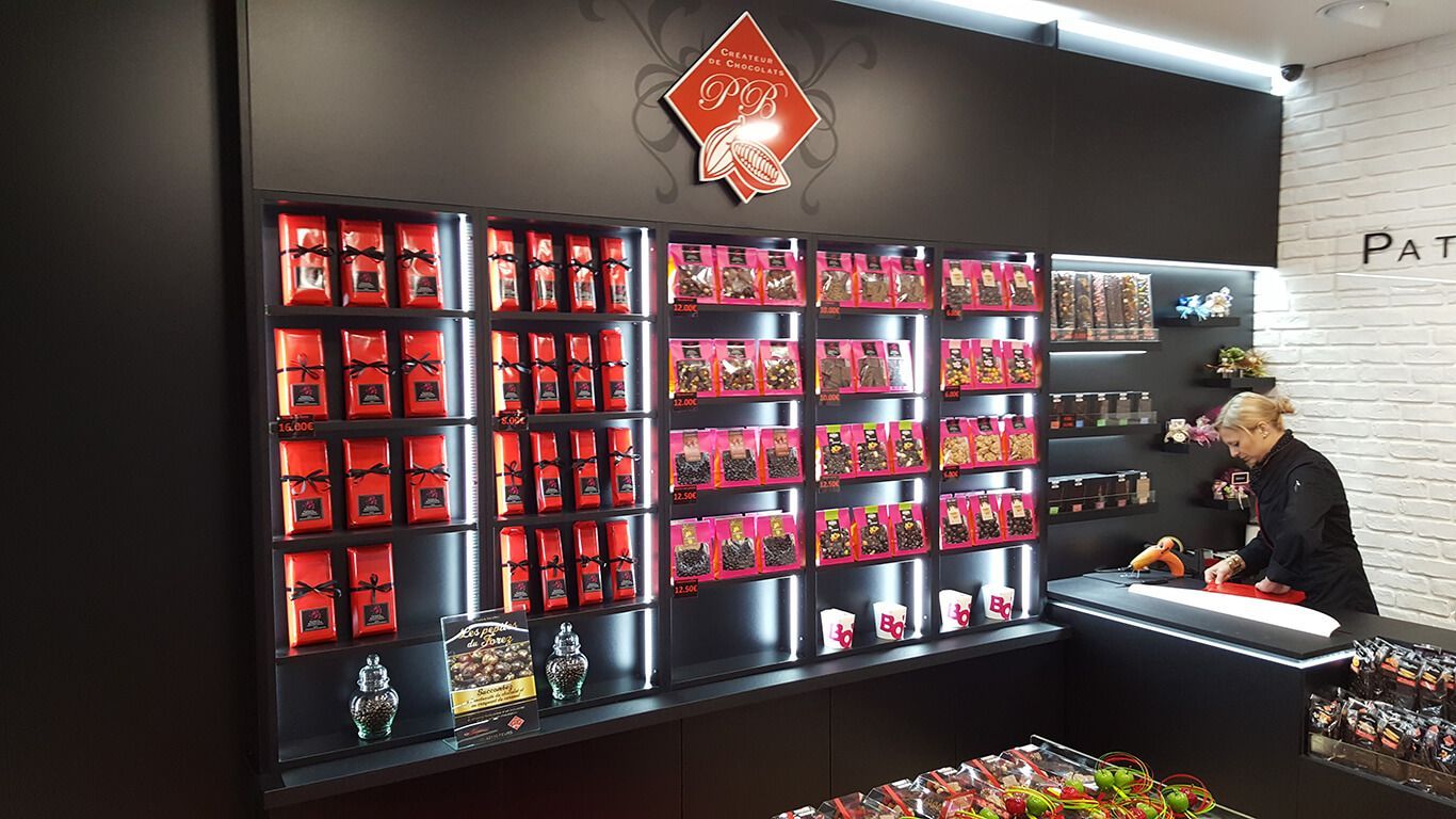 Intérieur d'un magasin présentant des rangées de boîtes de chocolats sur des étagères rétroéclairées, un employé travaillant au comptoir.