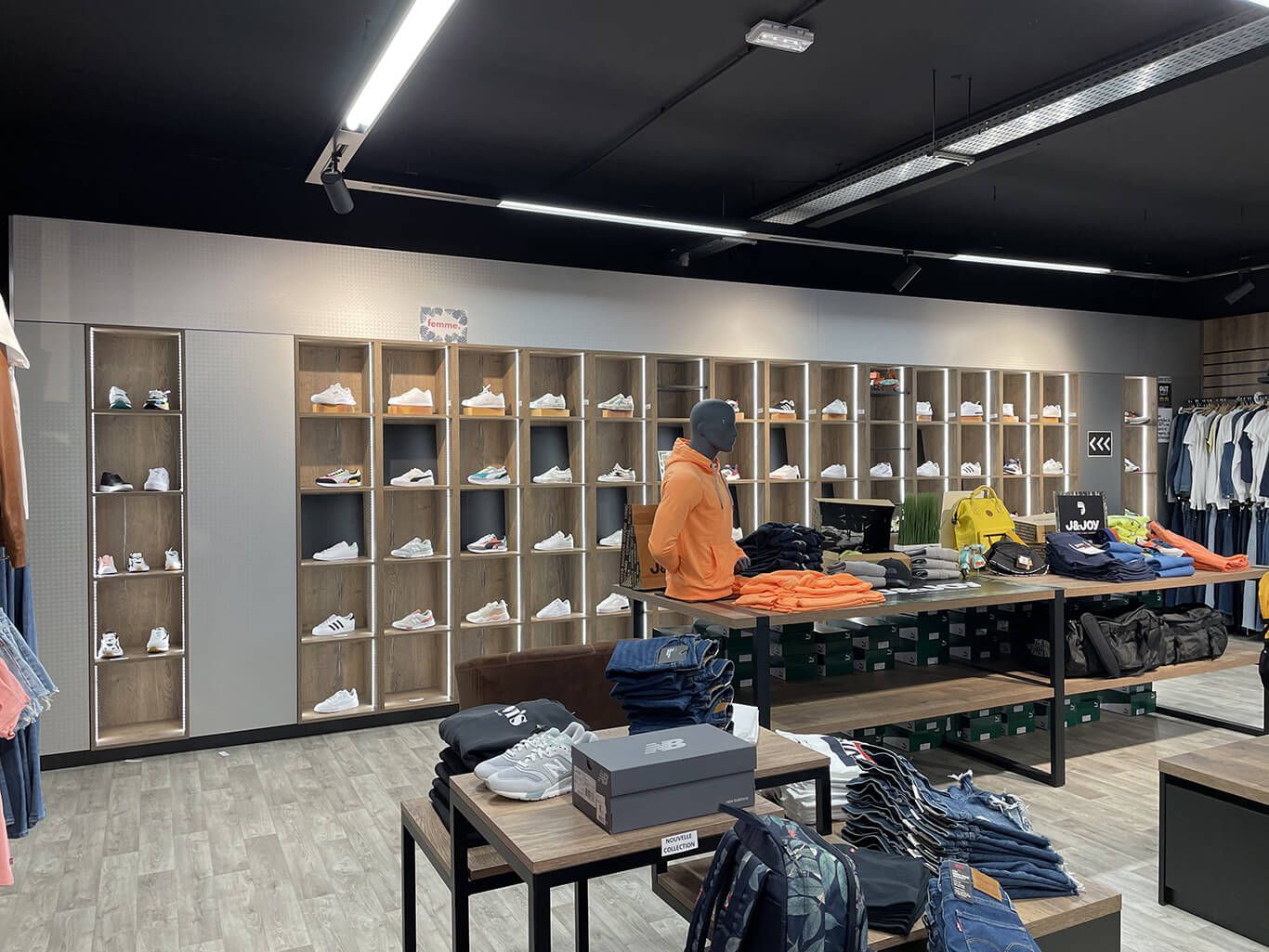 Intérieur d'un magasin de vêtements avec des étagères de baskets, des présentoirs de vêtements et une personne debout près d'une table.
