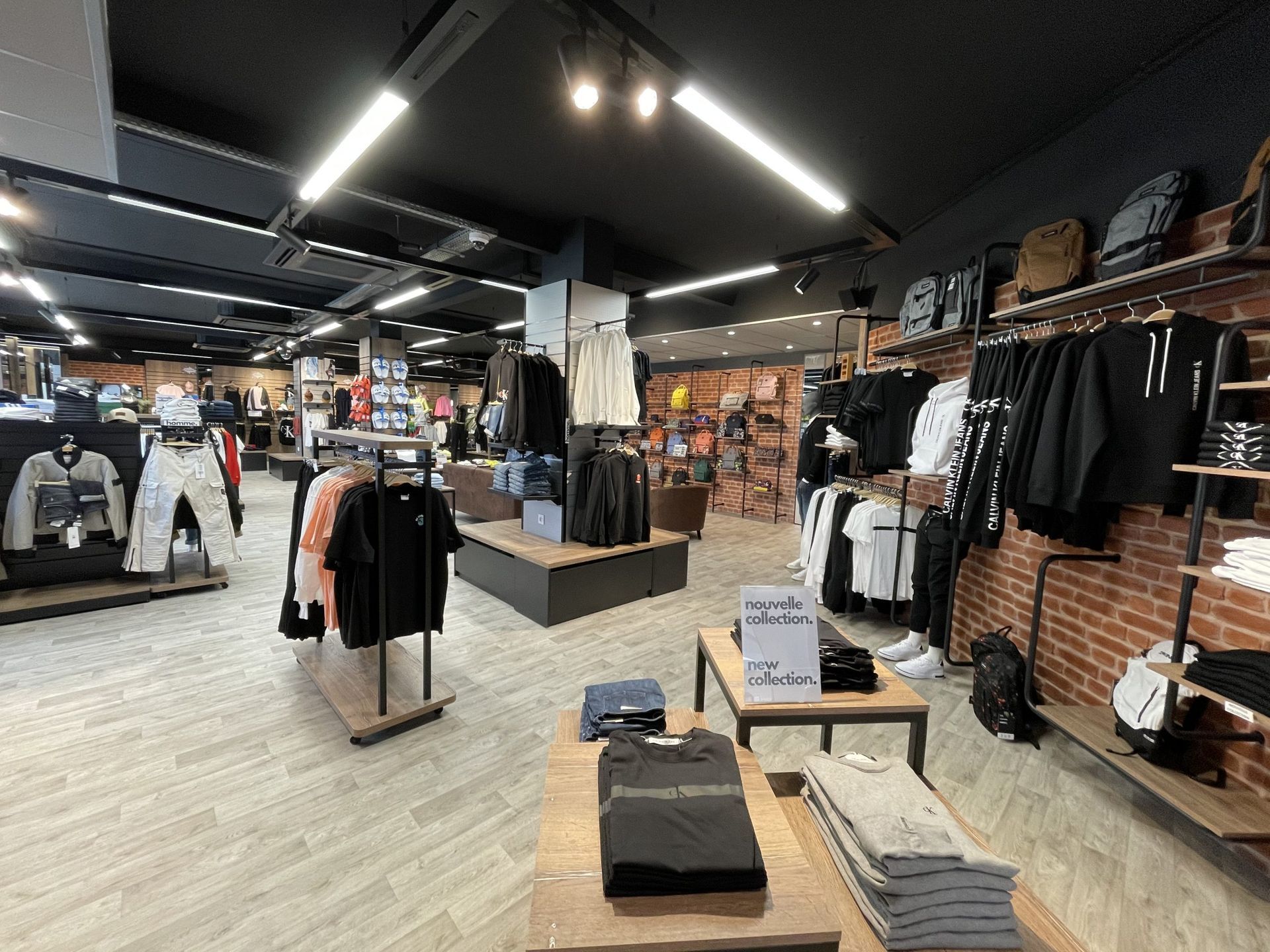 Intérieur d'un magasin de vêtements avec des portants, des chemises pliées sur des tables en bois et des murs en briques sous un éclairage vif.