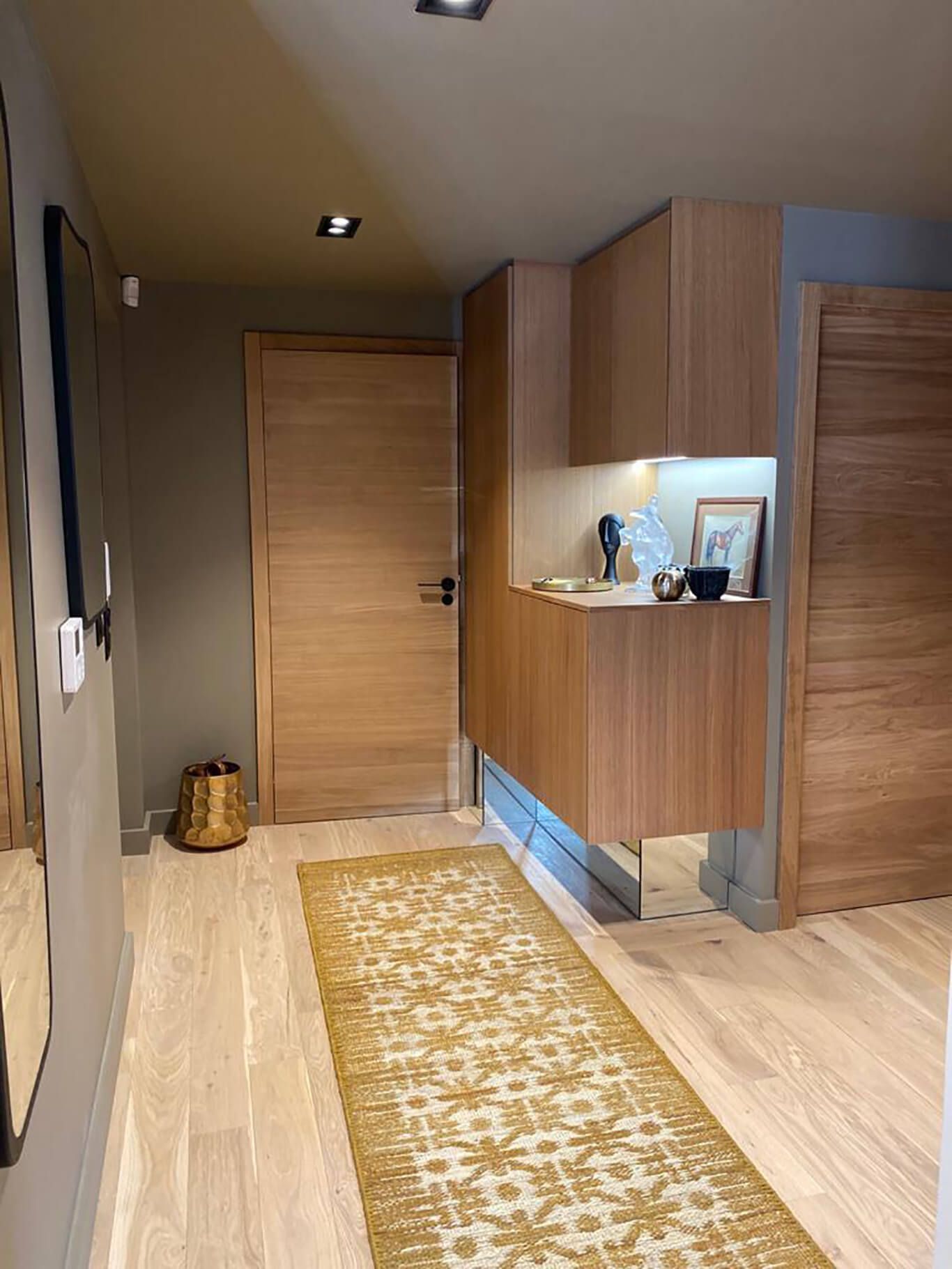 Un couloir moderne avec un parquet clair, un tapis jaune à motifs, des placards en bois et deux portes en bois fermées.