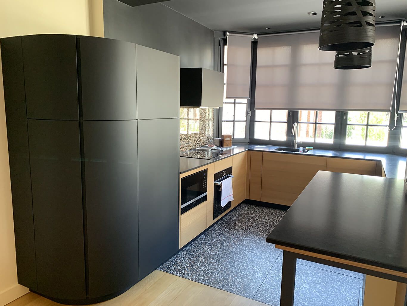 Une cuisine moderne avec des armoires incurvées noir mat, des meubles bas en bois clair, un îlot foncé et un sol moucheté.