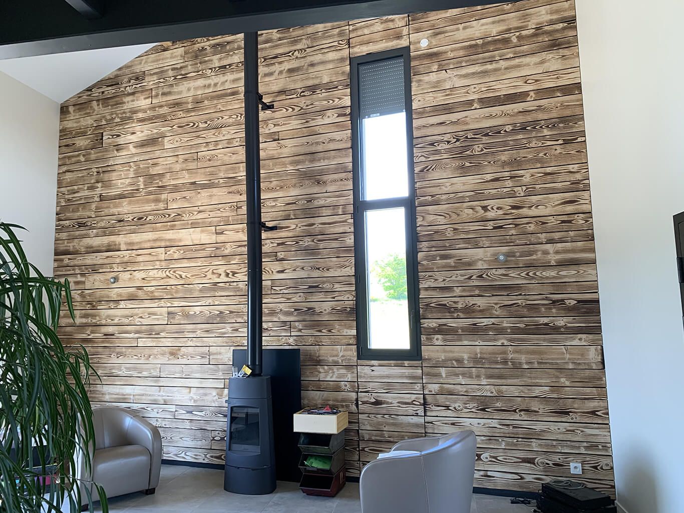 Une pièce avec un haut mur recouvert de planches de bois horizontales texturées, un poêle à bois noir et une fenêtre verticale.