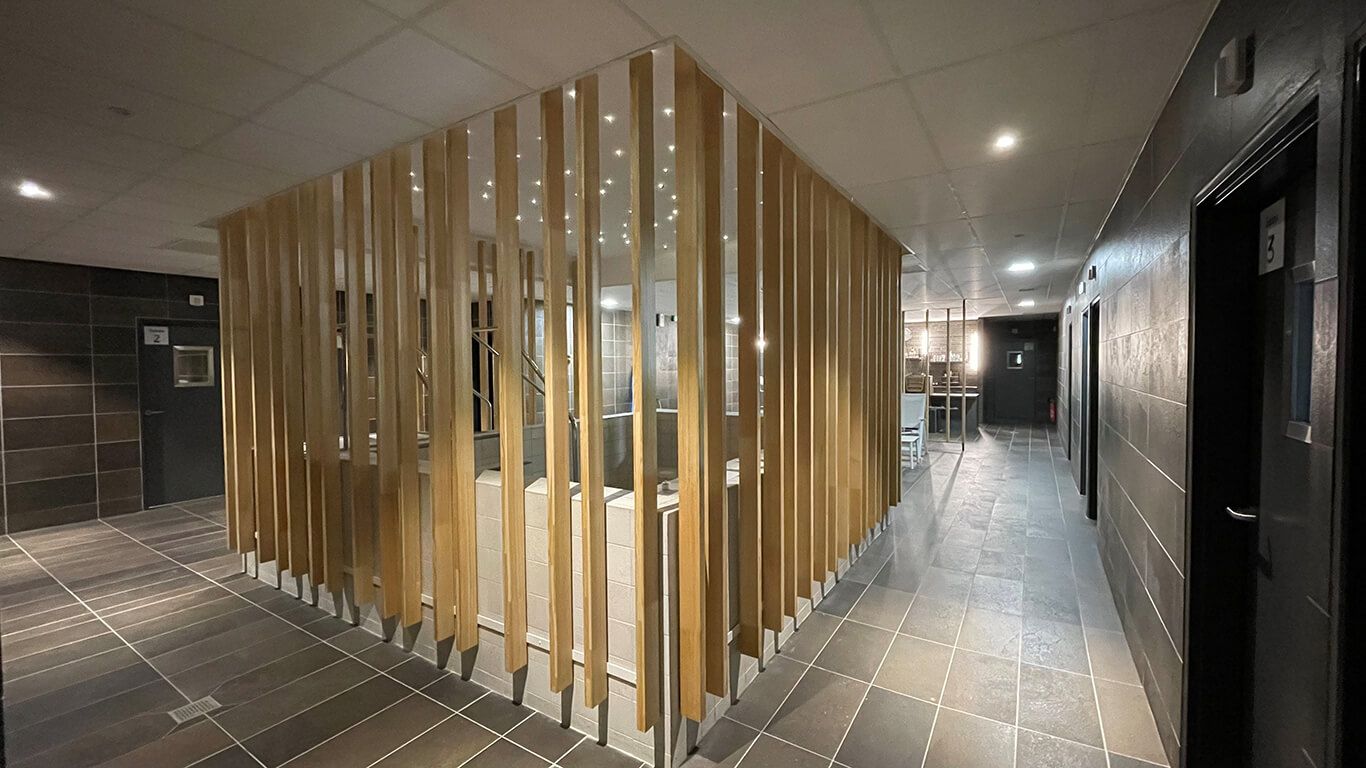 Un couloir avec un comptoir d'accueil moderne, délimité par des lattes de bois verticales sur un sol carrelé et éclairé par des spots encastrés.