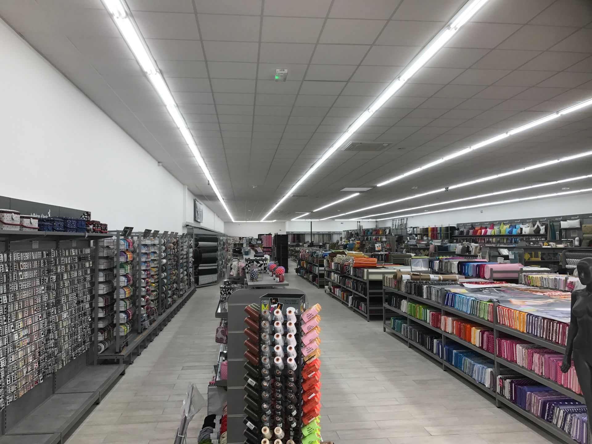Une longue allée lumineuse dans un magasin de tissus, avec des étagères remplies de rouleaux de tissu colorés de part et d'autre.