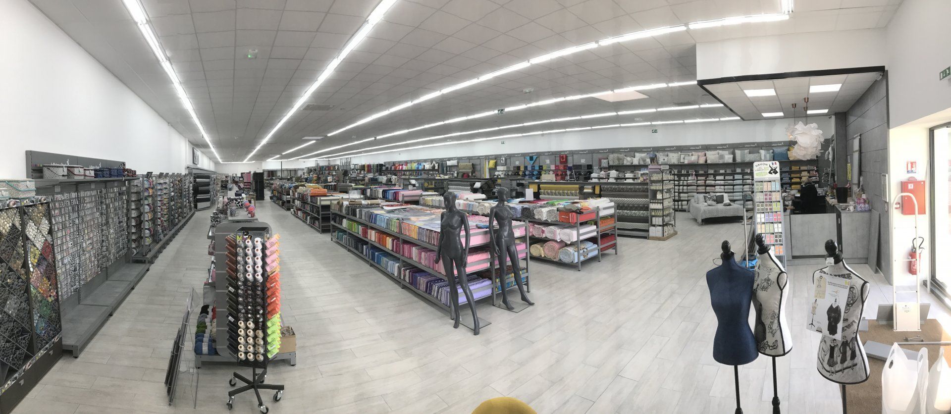 Vue grand angle d'un magasin de vêtements et de tissus avec des rangées d'étagères, des présentoirs et des mannequins sans tête.