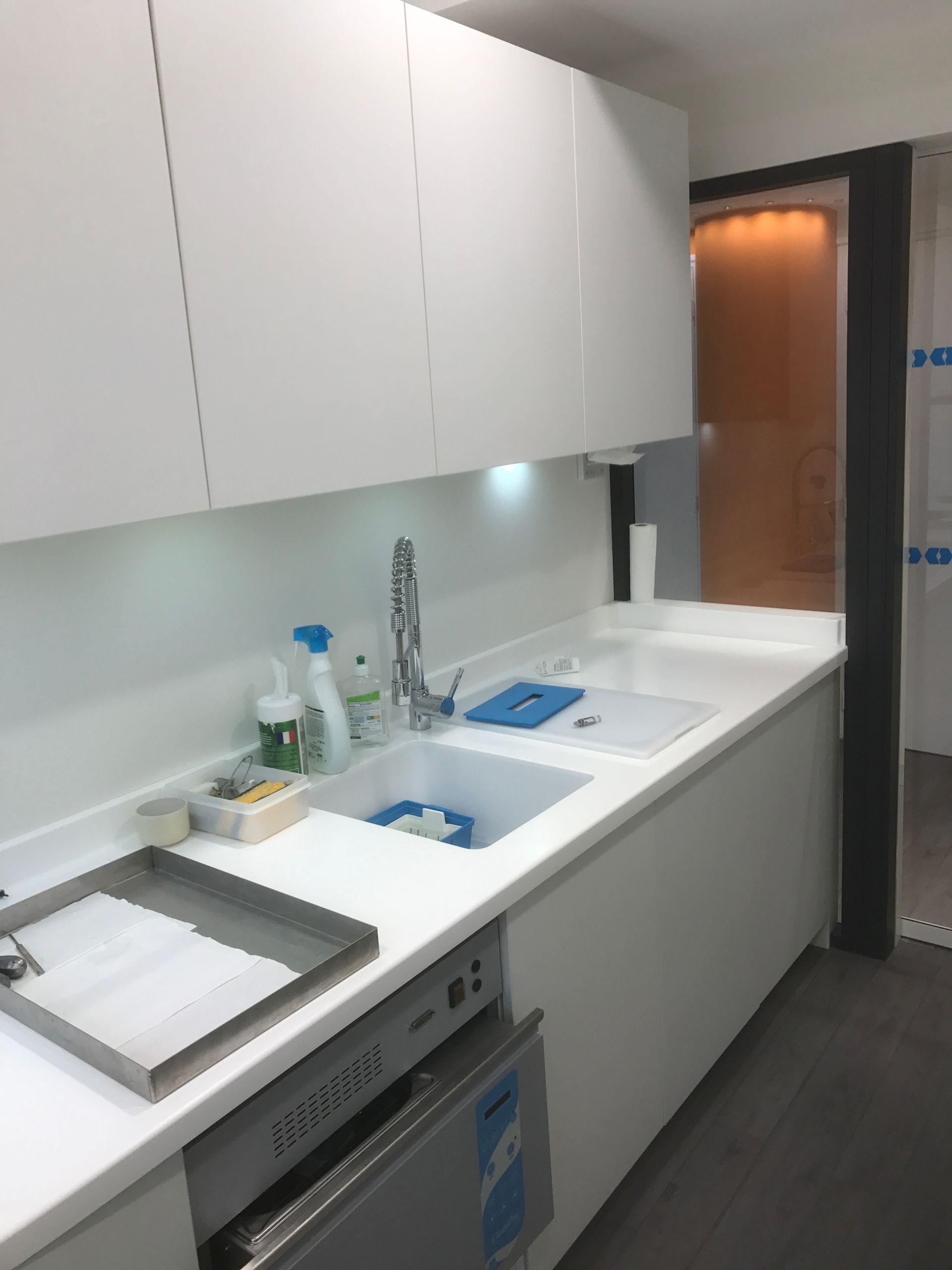 Un espace cuisine clinique moderne, blanc, avec un évier, des appareils électroménagers en acier inoxydable sous le comptoir et des armoires suspendues.