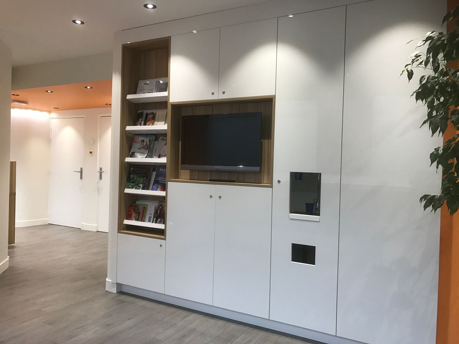 Meuble mural moderne avec placards blancs, niche TV encadrée de bois, étagères ouvertes et distributeur intégré dans un couloir lumineux.