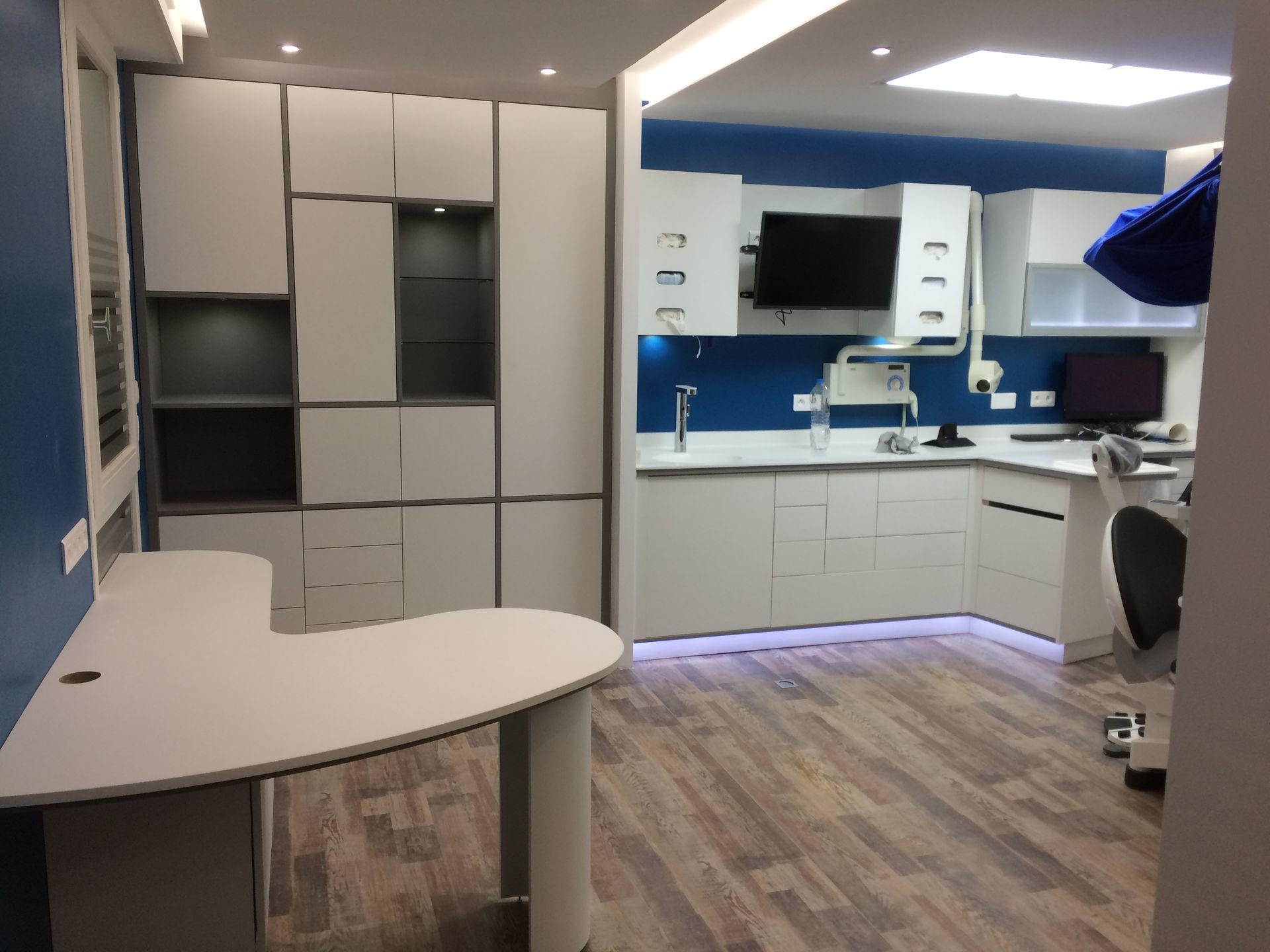 Un cabinet dentaire moderne et minimaliste avec des meubles blancs, un bureau incurvé, des murs d'accent bleus et un parquet clair.