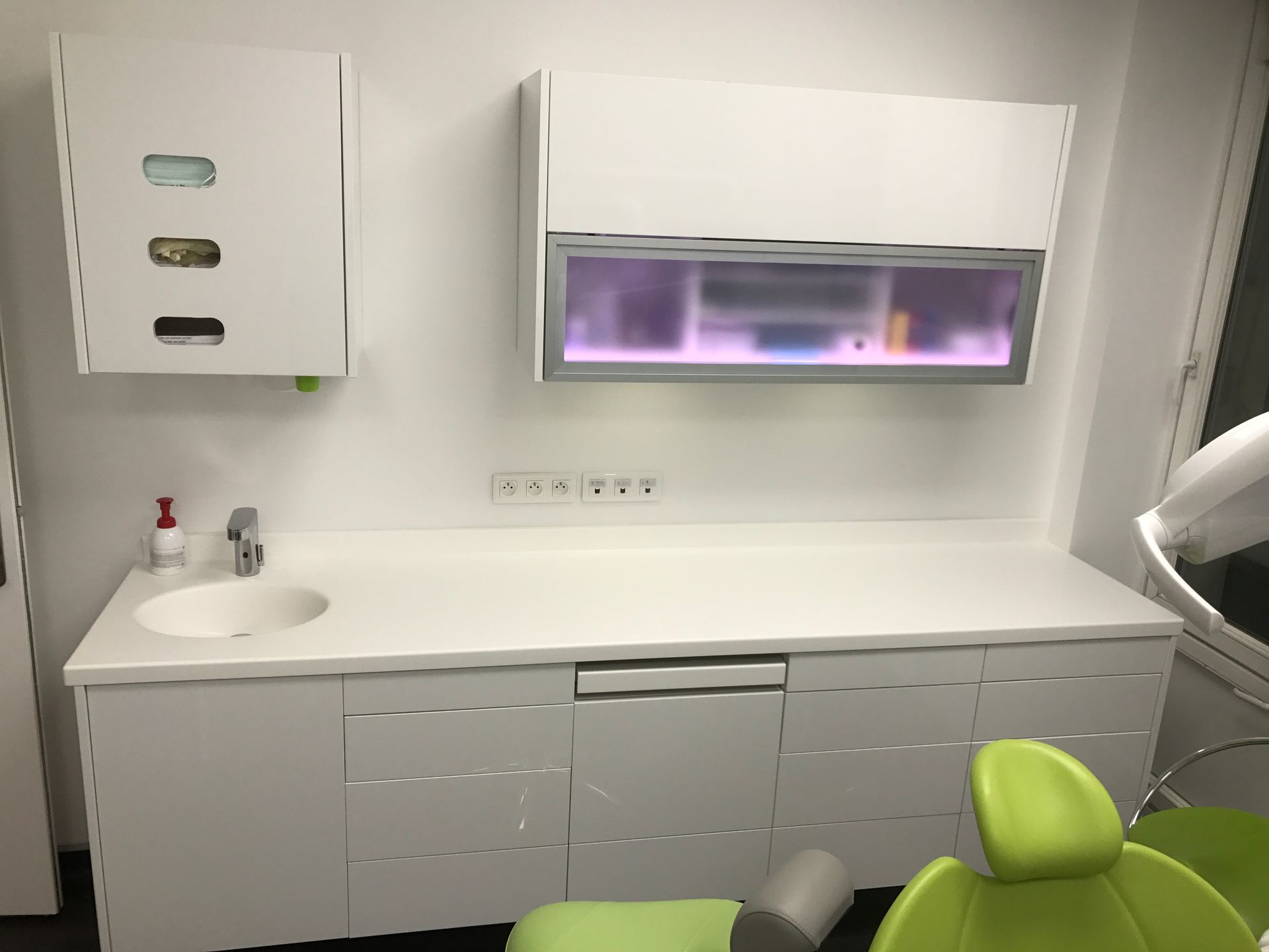 Une salle d'examen dentaire moderne avec un lavabo et des armoires blanches, des rangements muraux et une partie d'un fauteuil patient vert citron.