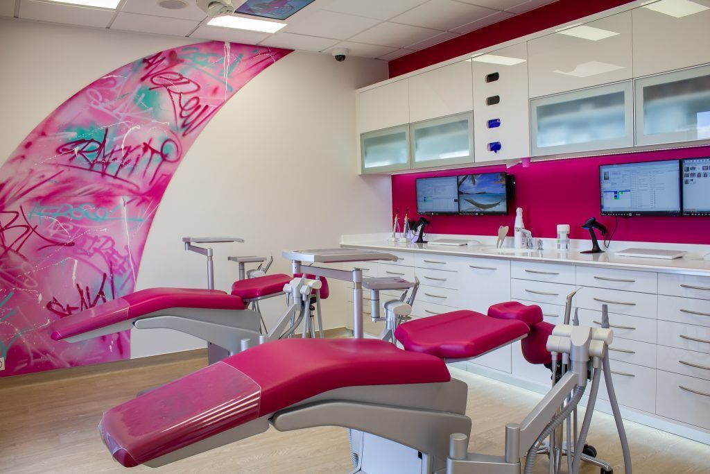 Cabinet dentaire moderne avec deux fauteuils roses pour les patients, des meubles blancs et une fresque murale artistique rose vif.