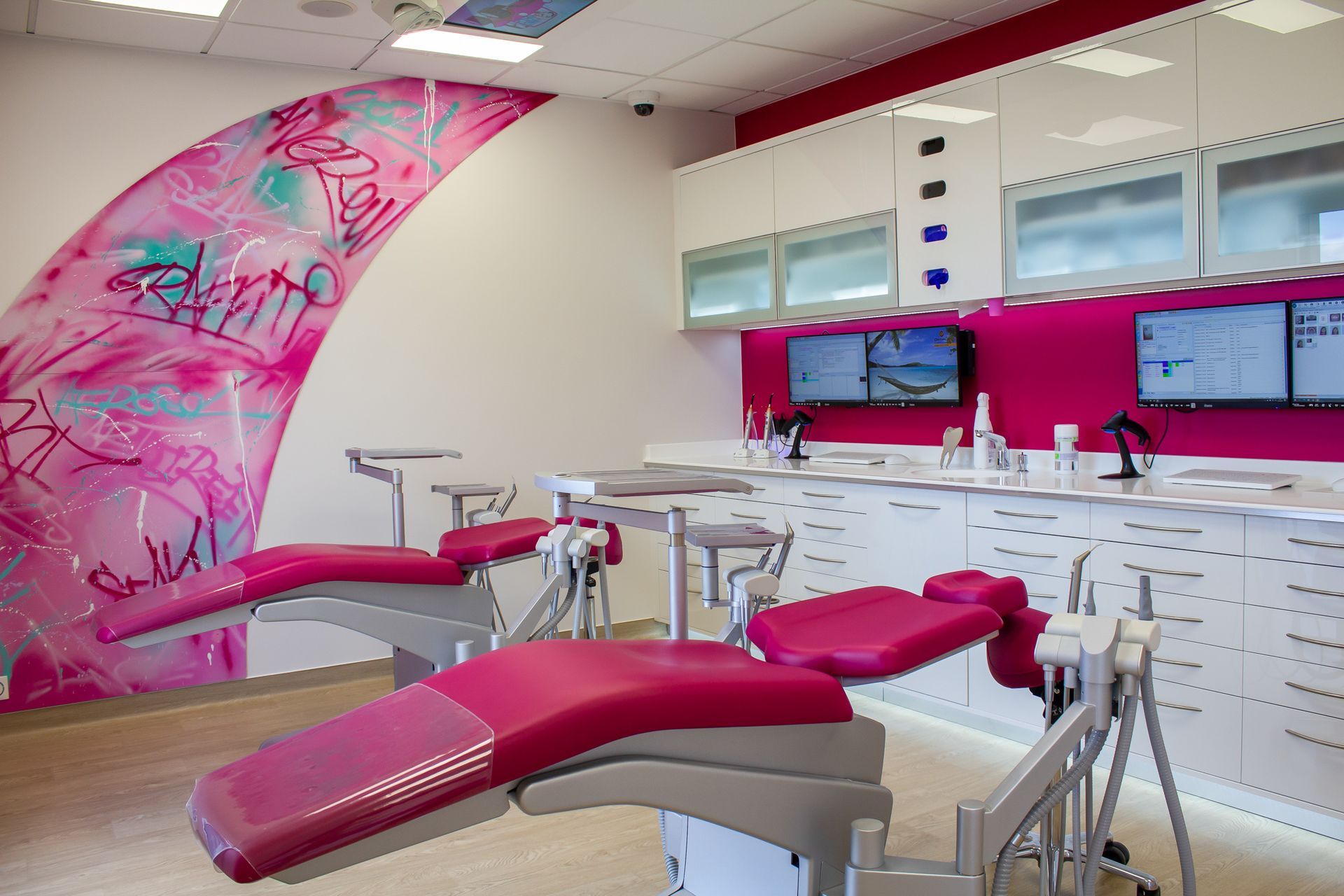 Un cabinet d'orthodontie moderne avec deux fauteuils dentaires roses, des armoires blanches et une fresque murale rose décorative.