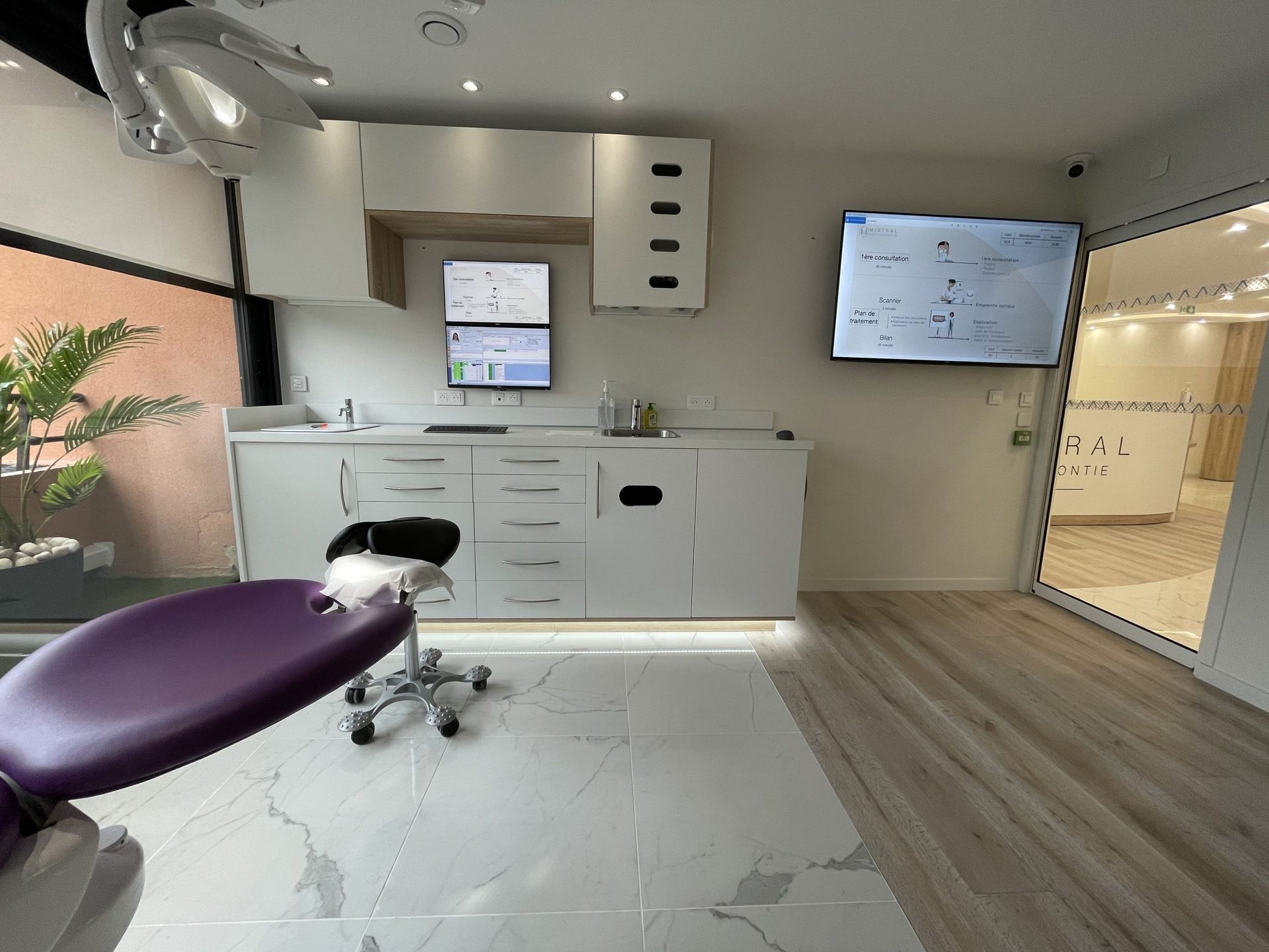 Une salle d'examen dentaire lumineuse et moderne, avec un fauteuil patient violet, des meubles blancs et des paravents muraux.
