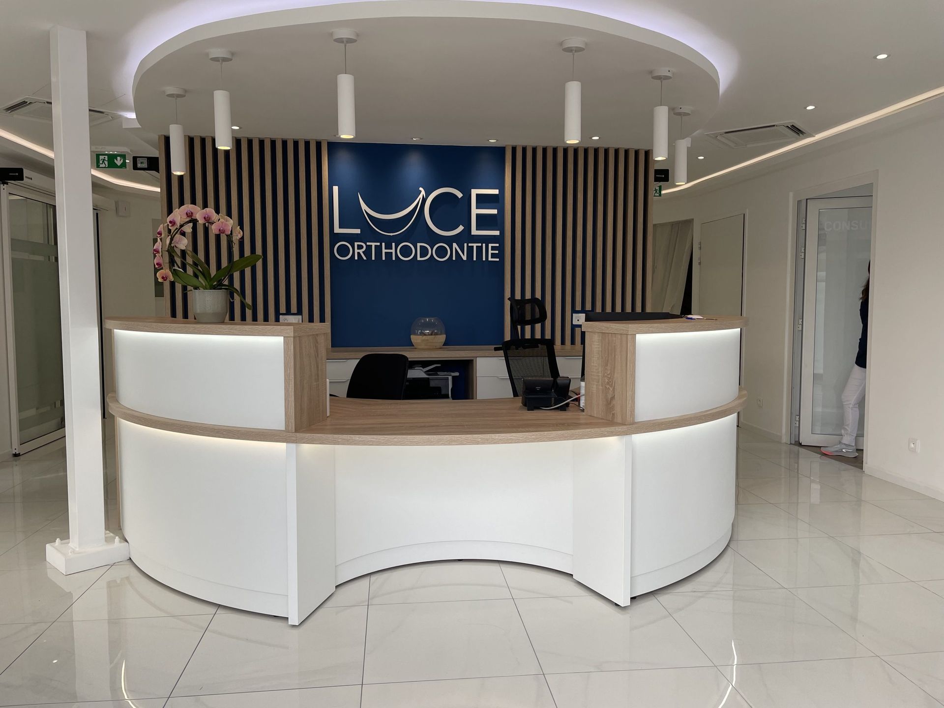 Espace d'accueil moderne avec un bureau blanc incurvé, le logo bleu de Luce Orthodontie sur un mur et un sol carrelé.