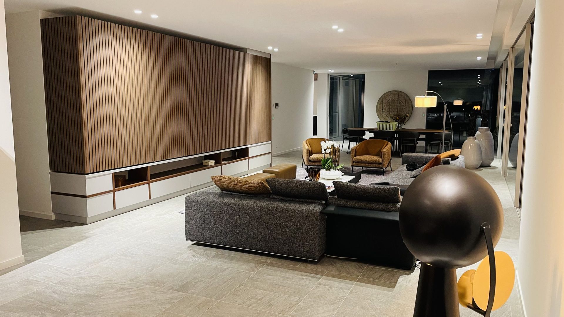 Un salon moderne avec un grand mur en bois à motifs, un canapé gris, des fauteuils en velours et une sculpture sphérique en façade.