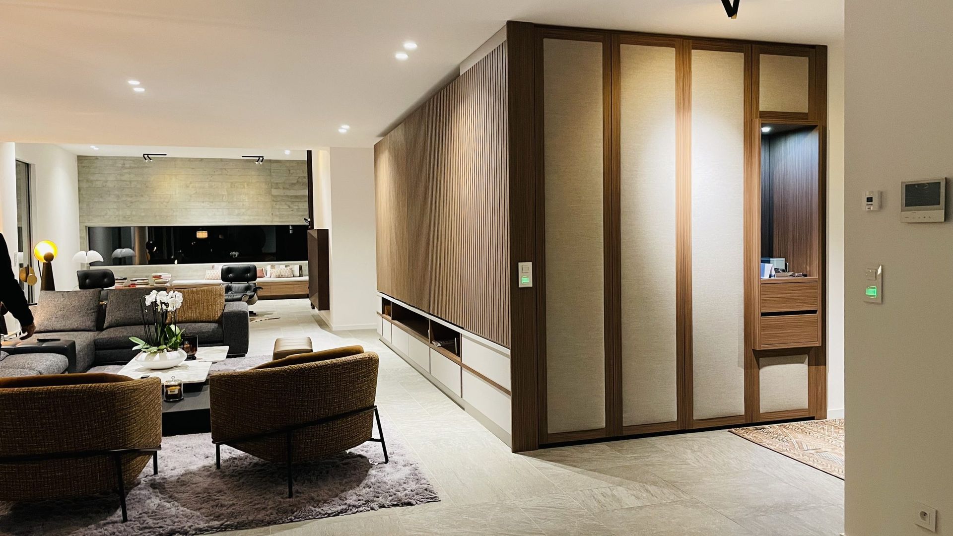 Salon moderne avec un canapé d'angle aux tons neutres, des chaises à motifs et un grand mur de rangement en bois allant du sol au plafond.