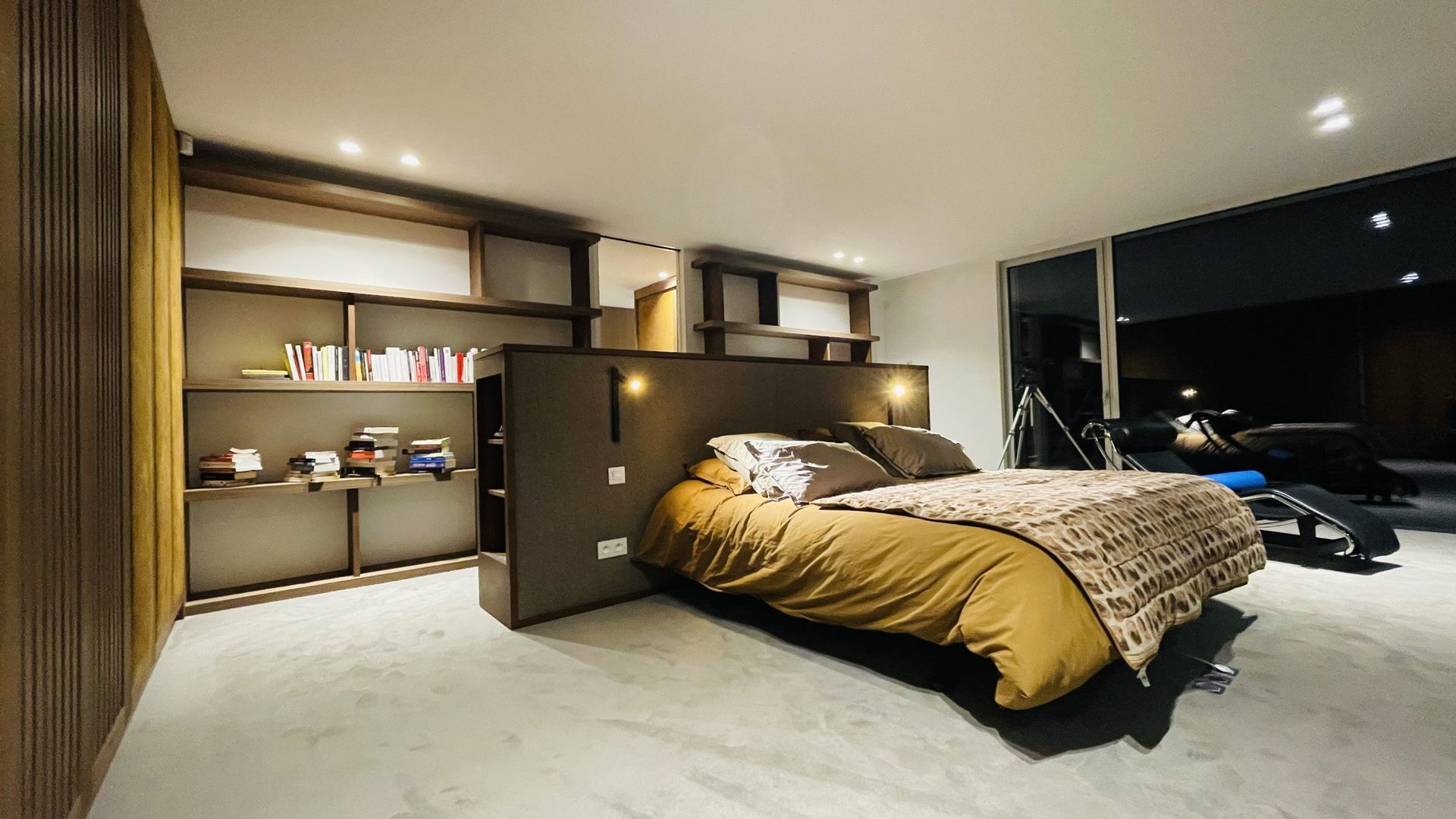 Une chambre moderne avec un lit central recouvert d'une couette beige, adossé à une grande étagère contre un mur sombre.