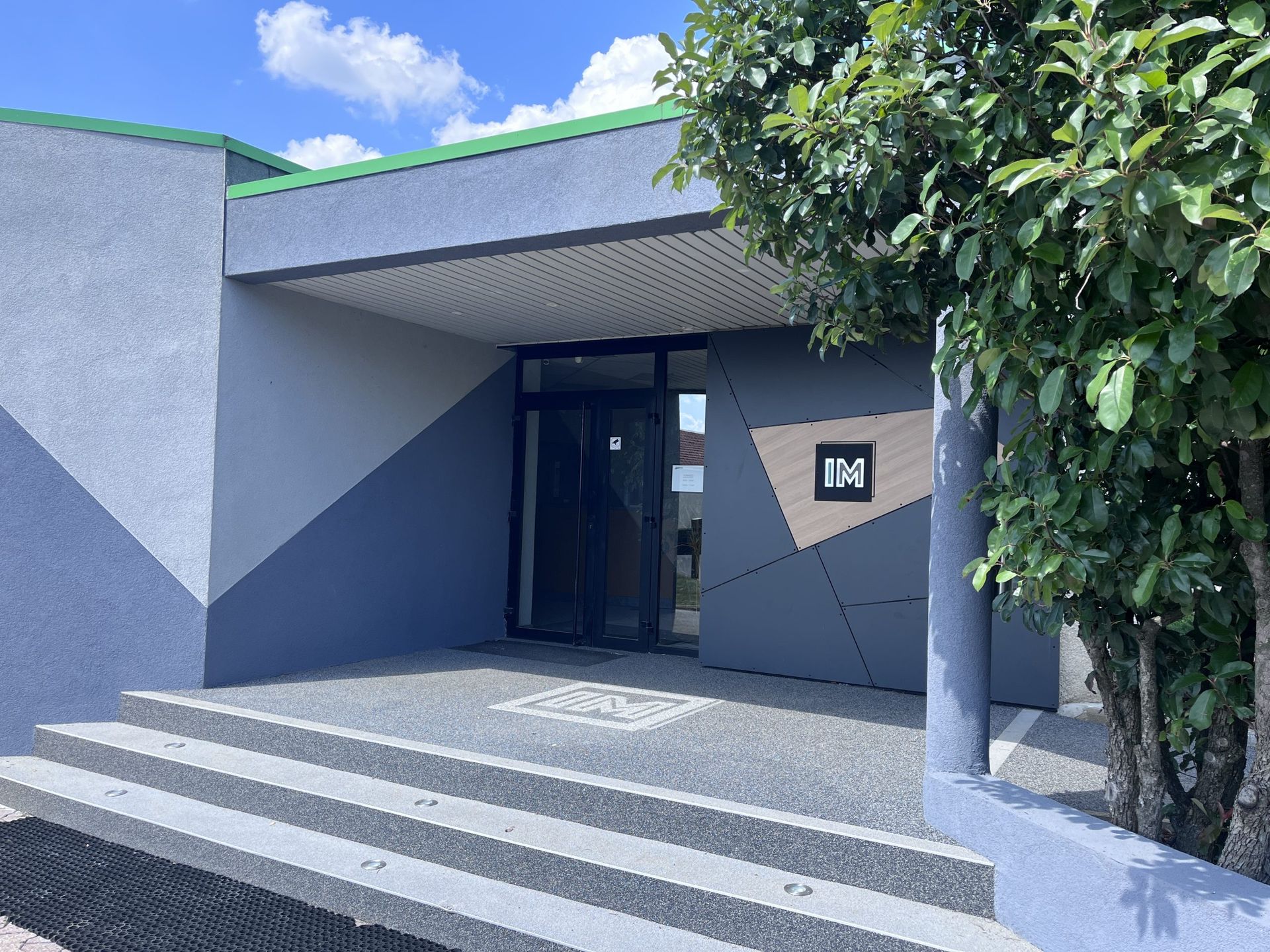 Entrée d'un bâtiment moderne avec des panneaux géométriques gris, un porche texturé et des marches menant à des portes vitrées sous un auvent.