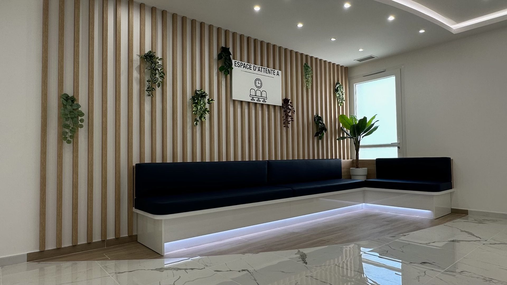 L'espace d'accueil comprend une banquette blanche en forme de L avec des coussins bleus, un mur en bois à claire-voie orné de plantes et une fenêtre lumineuse.