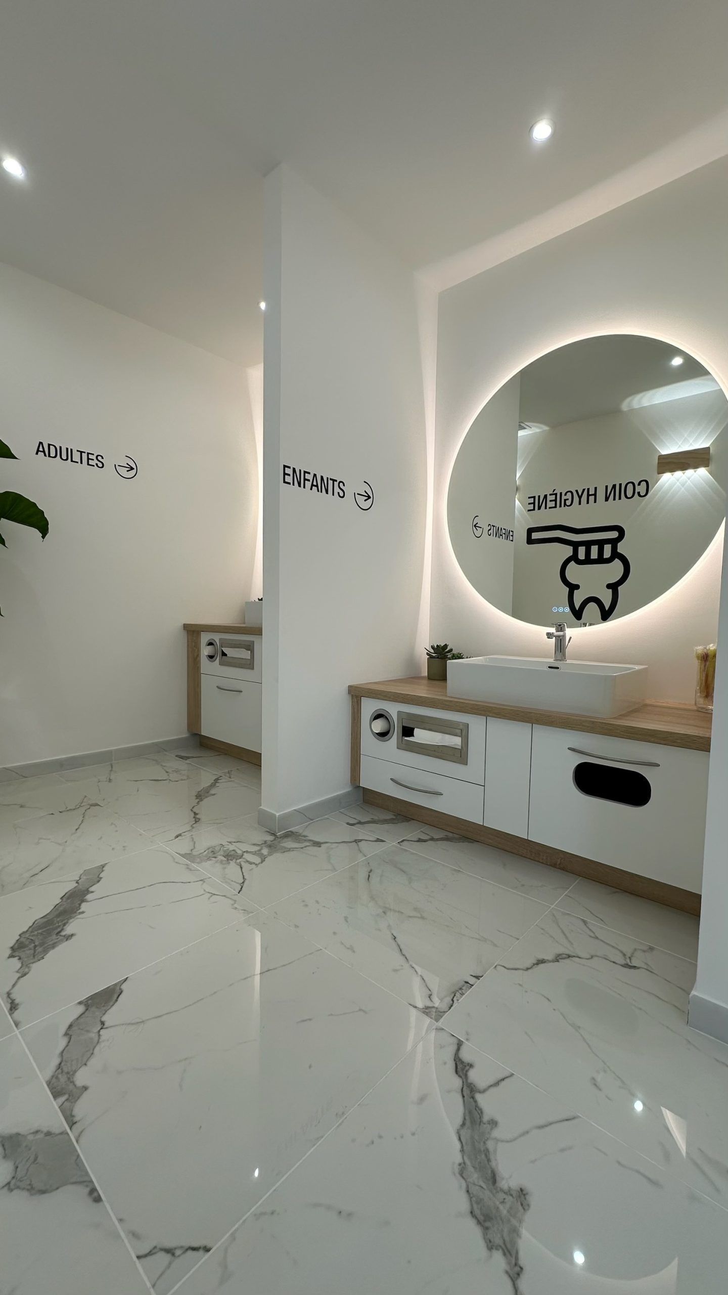 Toilettes modernes de clinique dentaire avec sol en marbre blanc, deux lavabos avec miroirs rétroéclairés et autocollants pour brosses à dents.