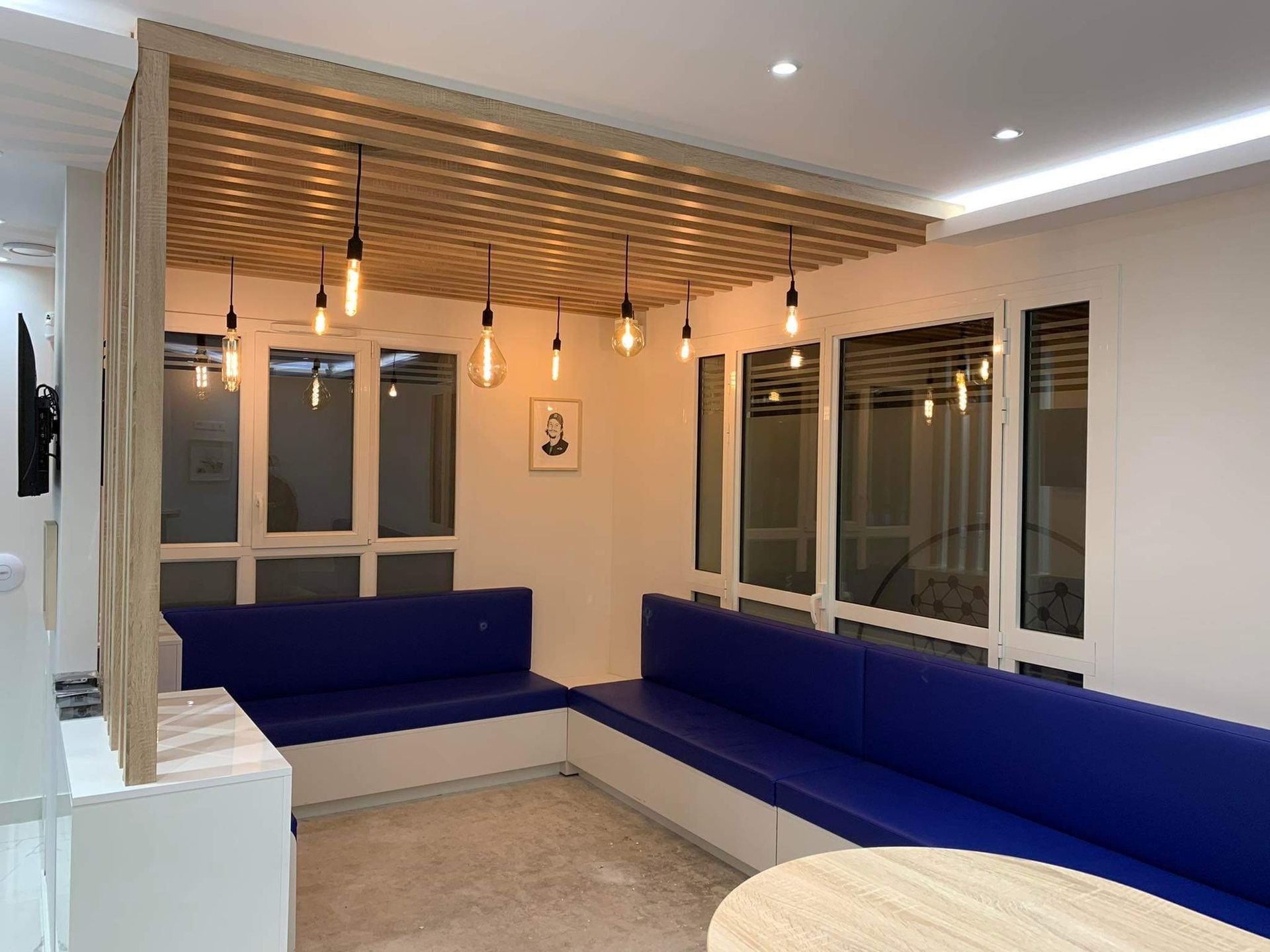 Salle d'attente moderne avec banquettes bleues rembourrées, plafond à lattes de bois et ampoules suspendues de style Edison.
