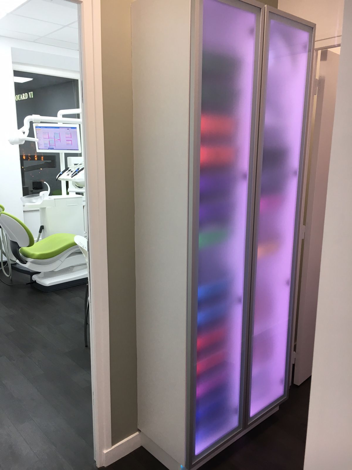Une haute armoire éclairée aux portes en verre dépoli violet se dresse à côté d'un cabinet dentaire où se trouve un fauteuil vert citron.