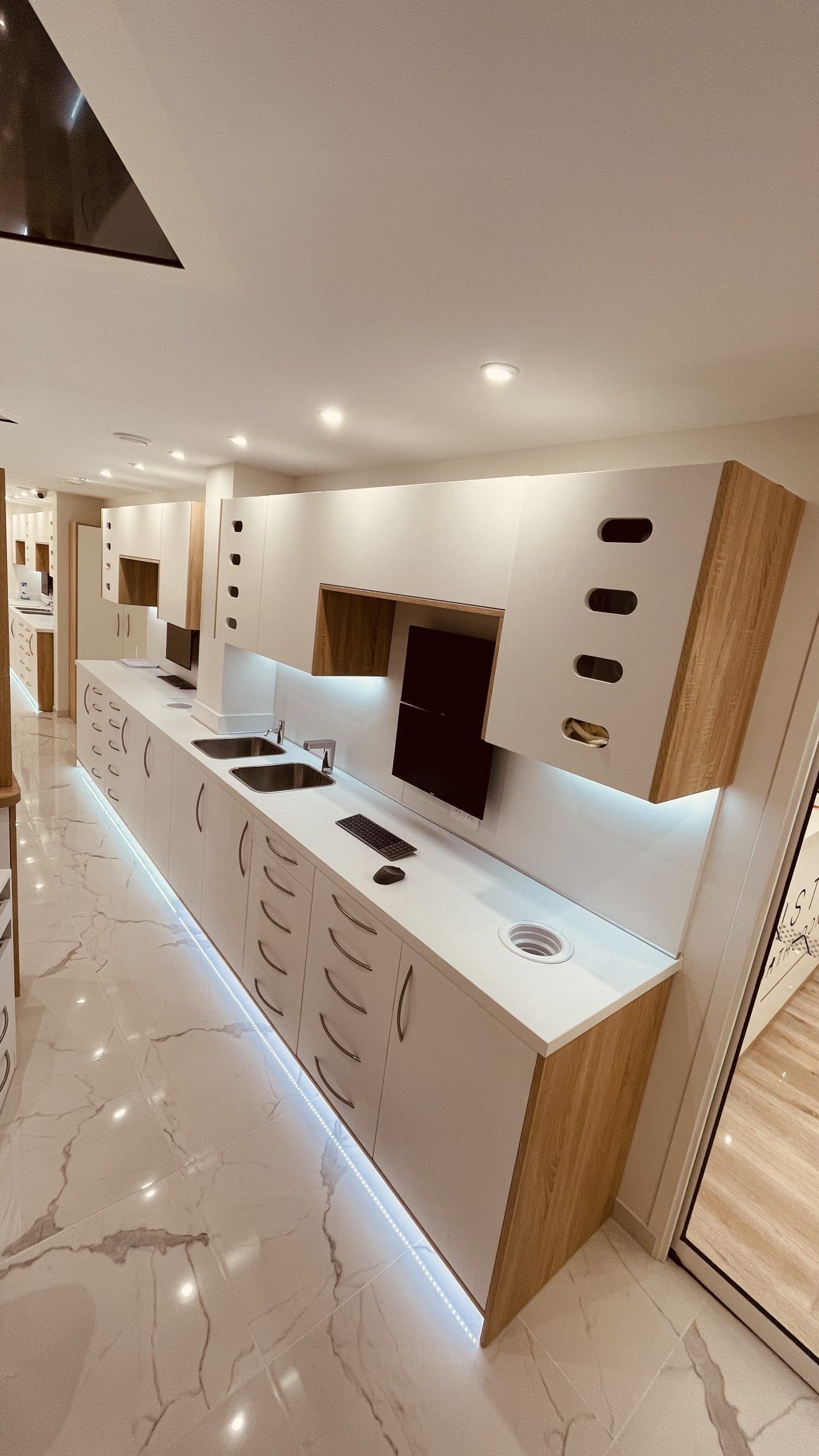 Intérieur moderne d'un cabinet dentaire avec des meubles blancs, des côtés en bois, un éclairage sous les meubles et un sol imitation marbre.
