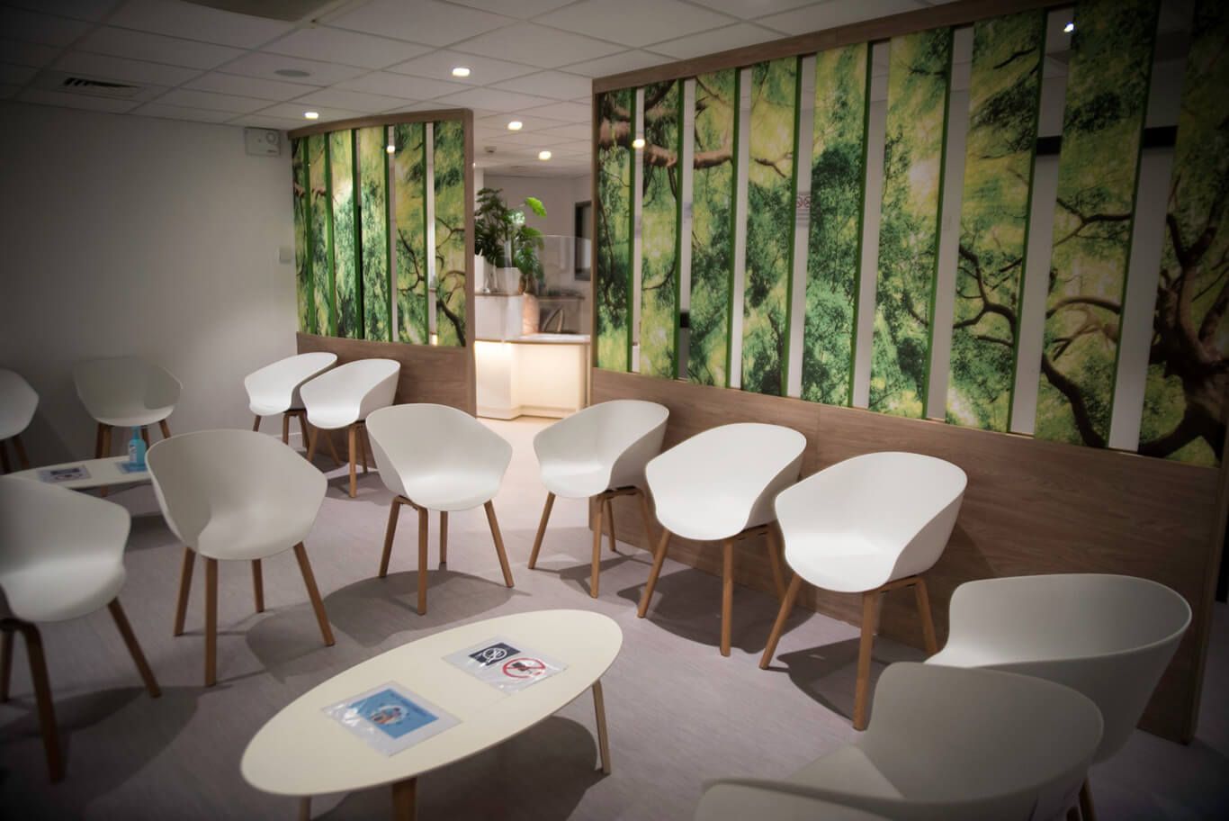 Une salle d'attente avec des chaises blanches disposées autour d'une table basse ovale, devant des murs ornés d'une fresque représentant une forêt.