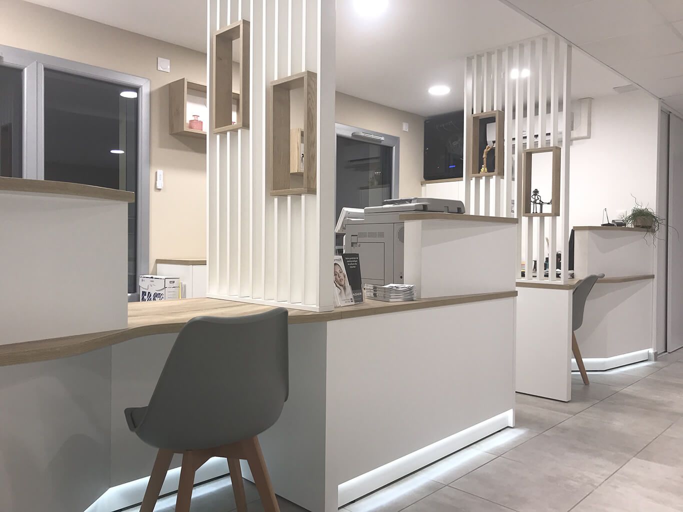 Espace d'accueil moderne avec bureaux blancs, touches de bois clair, chaises grises et éclairage LED discret au niveau du sol.