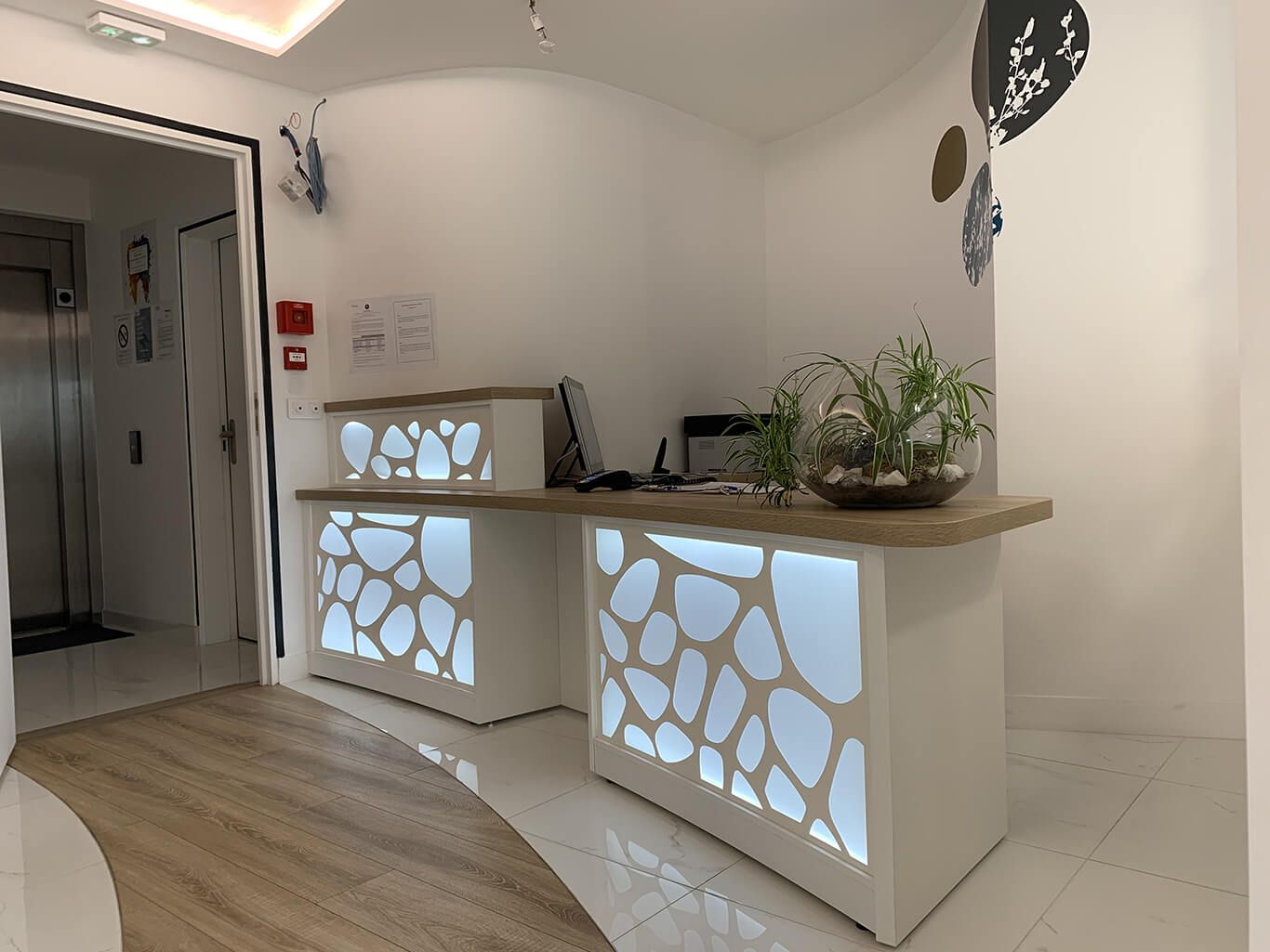 Dans un hall d'entrée, une réception moderne ornée de motifs organiques rétroéclairés, un comptoir en bois et une plante en pot.