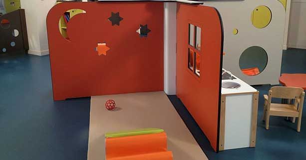 Une aire de jeux pour enfants comprenant des cloisons orange découpées en forme d'étoiles et de lune, une petite cuisine jouet et un ballon.