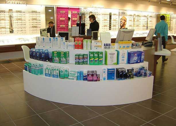 Dans un magasin d'optométrie, une étagère incurvée présente divers produits de soins oculaires, tandis que des employés travaillent à l'arrière.