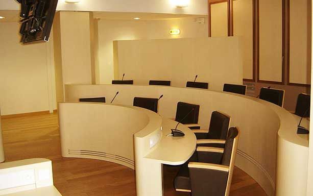 Une table de conférence ou de salle d'audience moderne et incurvée, avec des chaises et des microphones noirs, sur fond de murs couleur crème.