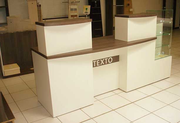 Un comptoir d'accueil en bois blanc et foncé avec une vitrine sur la droite et l'enseigne TEXTO au centre.