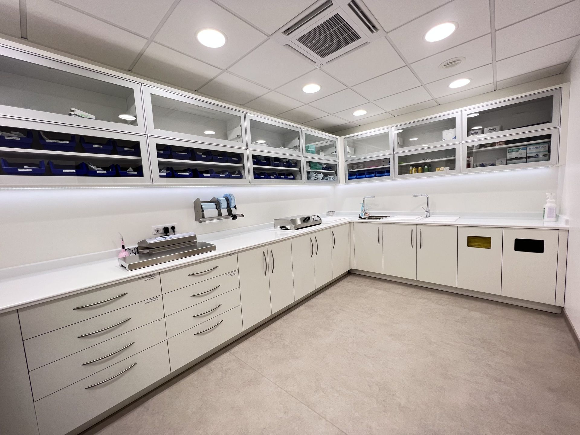 Une salle médicale ou un laboratoire moderne et lumineux, avec des armoires blanches, des rangements muraux vitrés et un double lavabo.