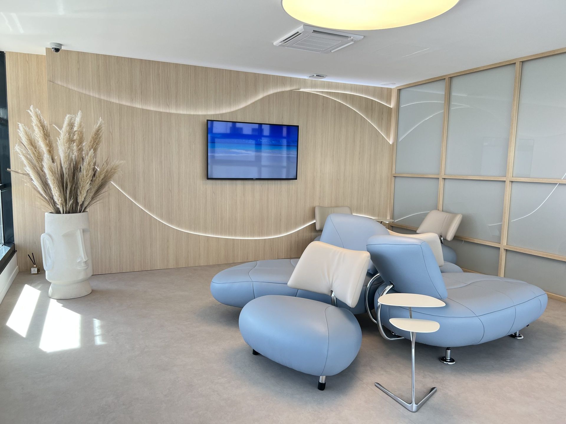 Un espace d'attente avec des canapés incurvés bleu clair, une table d'appoint blanche, une télévision sur un mur lambrissé et un grand vase au sol.