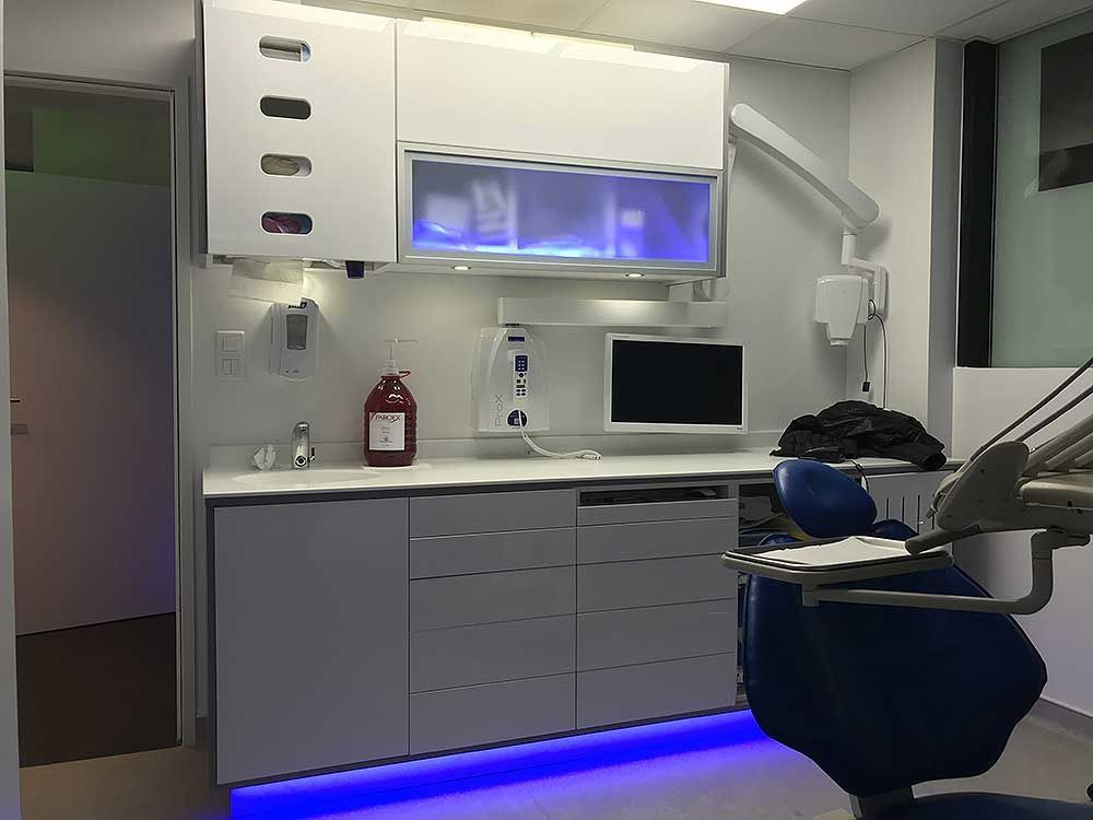 Une salle d'examen dentaire moderne avec éclairage LED bleu sous les meubles, un fauteuil dentaire et du matériel médical clinique.