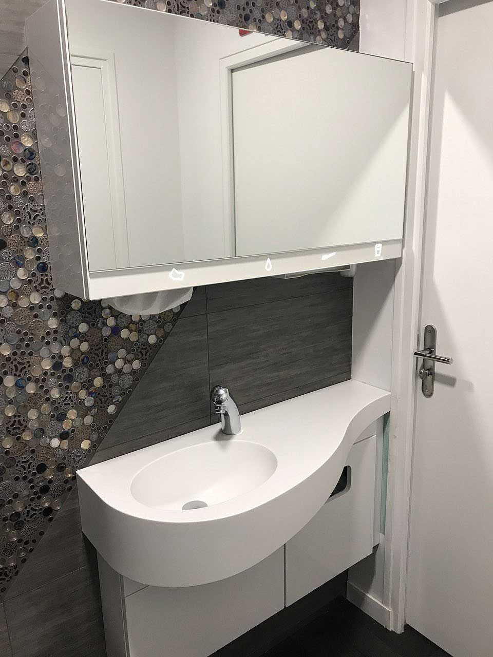 Un meuble-lavabo blanc moderne avec une vasque incurvée et une armoire à miroir, fixé contre un mur carrelé foncé et texturé.