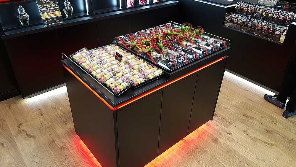 Un îlot de présentation dans un magasin, composé de plateaux de petits articles emballés, éclairés par un éclairage LED orange au niveau de la base.