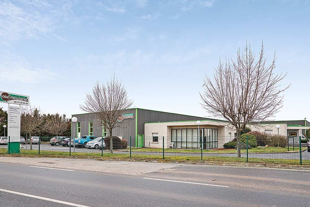 Un bâtiment commercial gris avec des touches de vert, un parking et des arbres dénudés derrière une clôture en grillage, sous un ciel bleu.