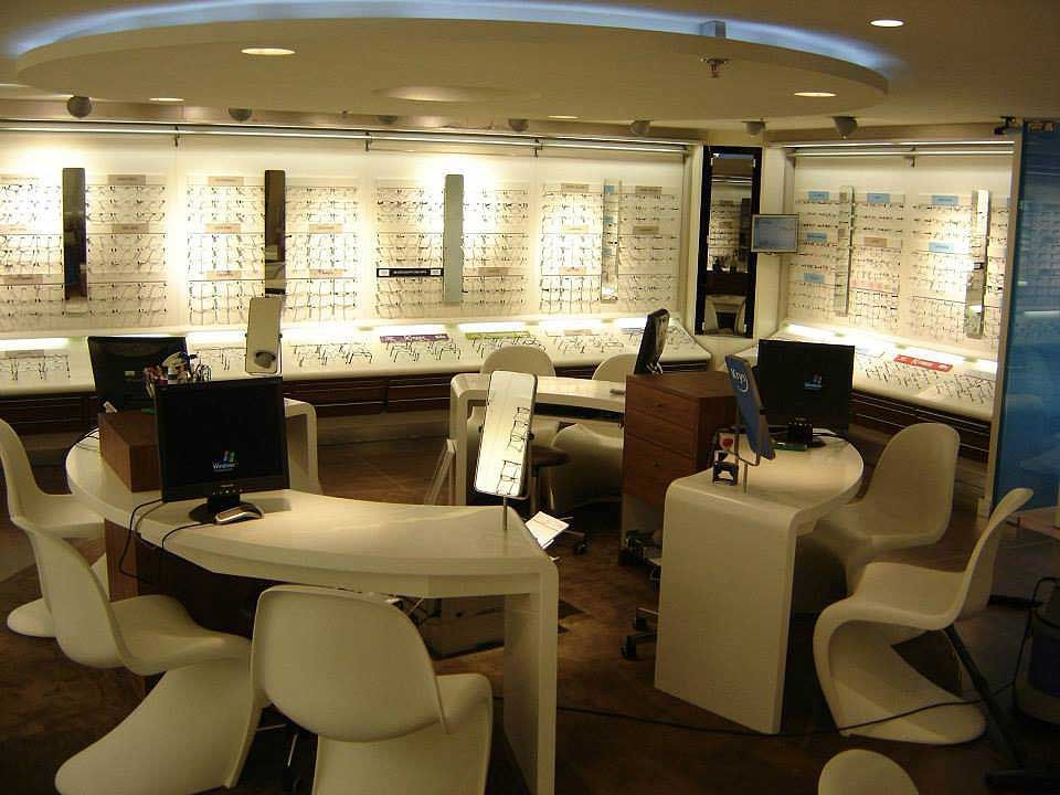 Une boutique d'optique moderne et lumineuse, avec des bureaux et des chaises blanches incurvées, et des présentoirs muraux lumineux remplis de lunettes.