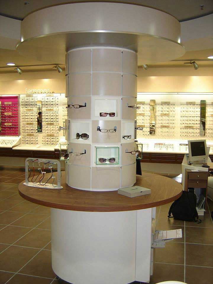 Un présentoir cylindrique contenant plusieurs paires de lunettes, placé au centre d'un magasin d'optique baigné de lumière.