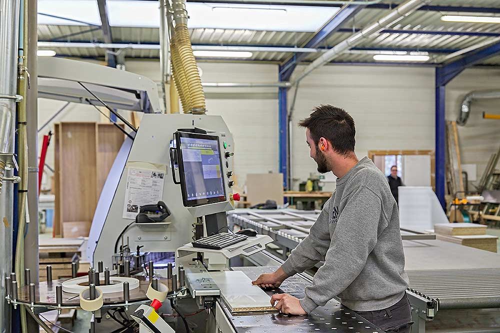 Un technicien manipule une plaqueuse de chants industrielle dans une usine de menuiserie baignée de lumière.