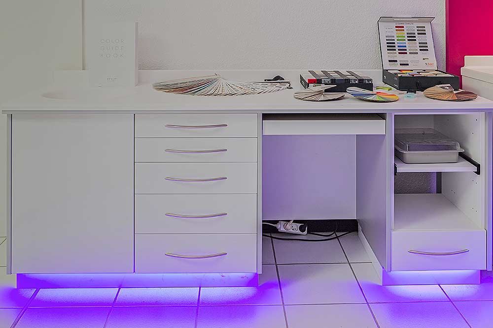 Un bureau blanc moderne avec plusieurs tiroirs, une étagère ouverte et un éclairage LED violet sous les meubles, sur un sol carrelé.
