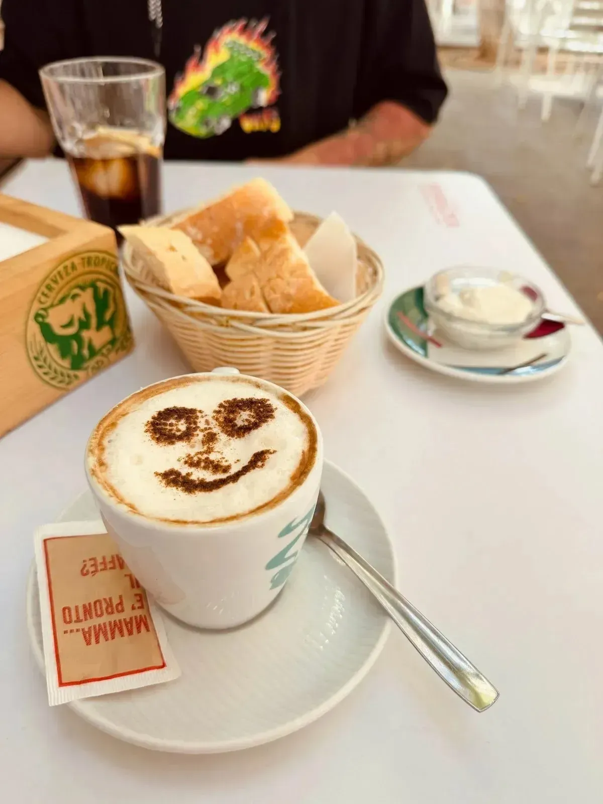 Café con una carita sonriente en la espuma, servido con pan y té en una mesa de café.